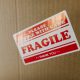 A closeup of a cardboard box with a red and white label that says, "Please handle with care. Fragile. Thank you."