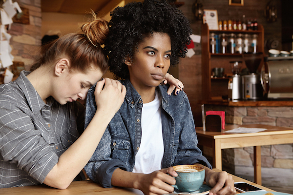 Young Caucasian female sitting at cafe, saying sorry, trying to apologize to her mad and angry African girlfriend after dispute or quarrel, begging for forgiveness. Disagreement in relationships