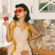 A woman poses on a kitchen counter in a shimmery dress wearing tensil around her shoulders and red sunglasses.