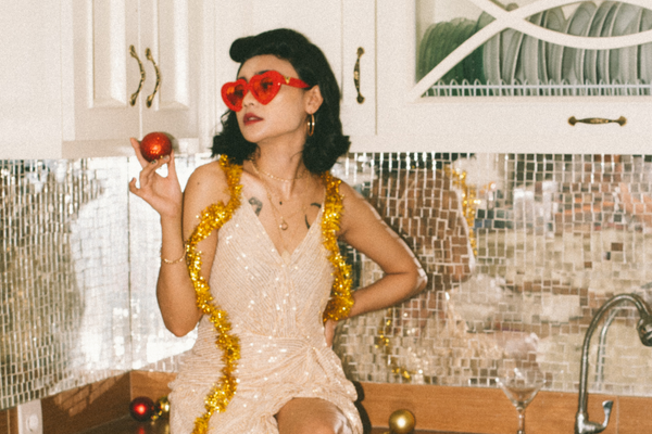 A woman poses on a kitchen counter in a shimmery dress wearing tensil around her shoulders and red sunglasses.