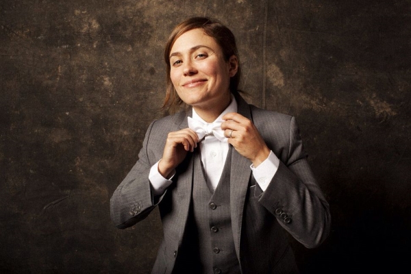A queer white woman wears a dark gray suit adjusts her white bow tie