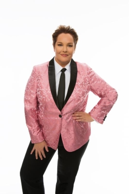 A masculine white woman wears a pink brocade blazer with black trim, black pants, a white button-up shirt, and a black tie. 
