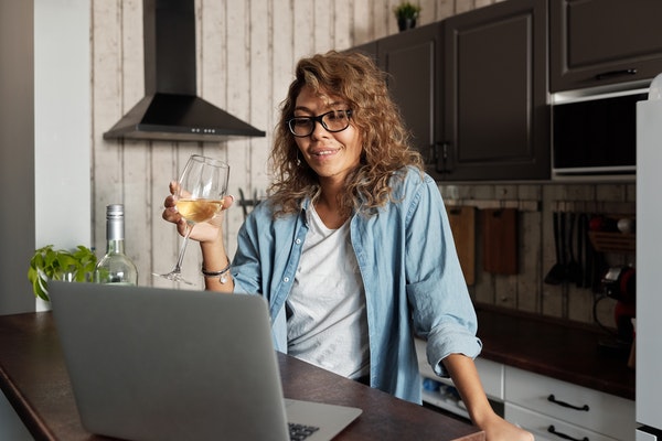 A woman holds a glass of white wine while standing in her kitchen. She's looking at a laptop that's sitting on her island.