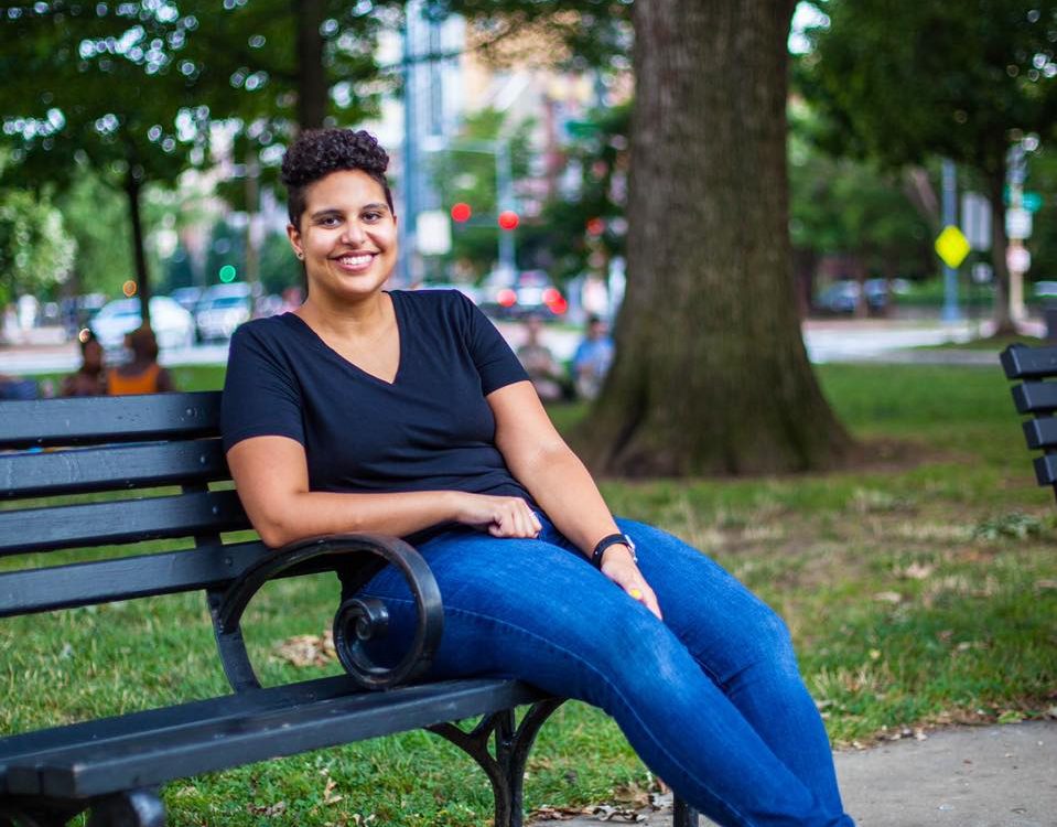 Rehana Mohammed, a Black bisexual Muslim woman with short black hair, sits on a park bench. She is wearing a dark blue v-neck t-shirt, blue jeans, and black and white sneakers.