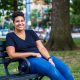 Rehana Mohammed, a Black bisexual Muslim woman with short black hair, sits on a park bench. She is wearing a dark blue v-neck t-shirt, blue jeans, and black and white sneakers.
