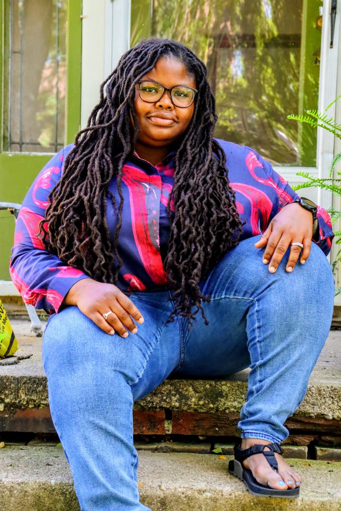 A Black person in a purple and pink top and jeans sitting