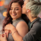 A woman hugs her girlfriend from behind while kissing her temple affectionately.