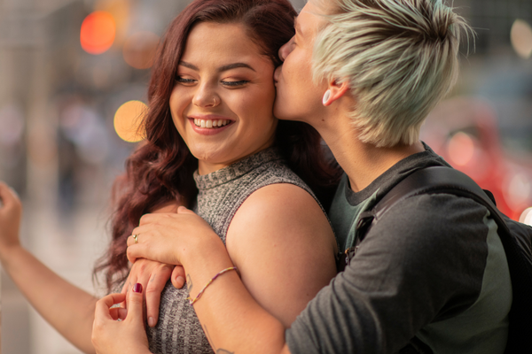 A woman hugs her girlfriend from behind while kissing her temple affectionately.