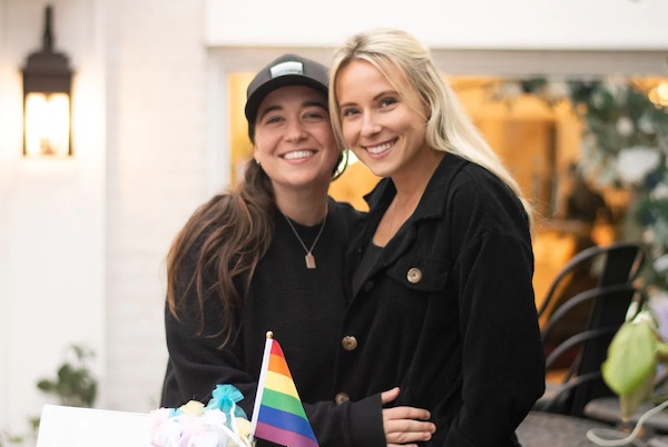 Queer wellness and mental health professionals Keely Antonio and Dani Max pose in front of a rainbow flag