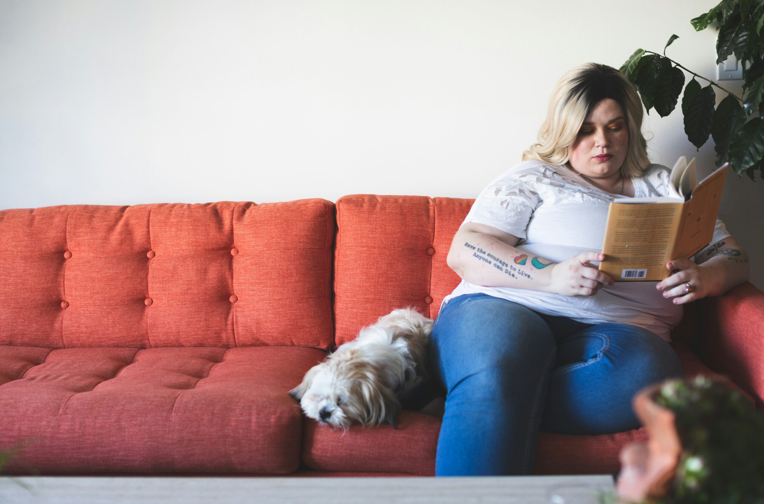A blonde woman reads a book on her red couch with her white dog at her side.