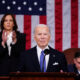 President Joe Biden stands in front of a microphone at a lecturn wearning a blue tie. Behind him, Vice President Kamala Harris stands clapping. In the background hangs a large American flag.