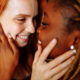 Two women hold each others' faces and smile at one another. One woman is white with red hair and the other is Black and has braids. Both look happy and in love.