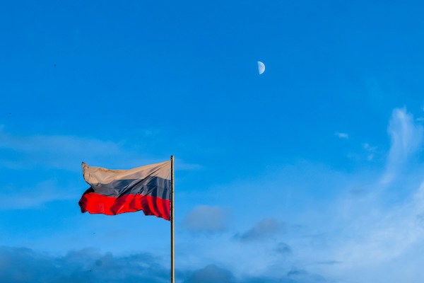 The Russian flag flies through the sky during the daylight. The moon is faintly visible in the background. The flag is tricolor: white on top, blue in the middle, red on bottom.