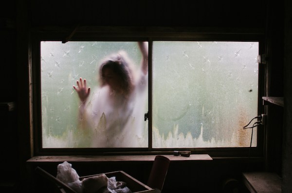 A figure is pressed up against a frosted window as if they ran into it. Their hands grasp at the window. In the foreground is a decrepit wooden room.