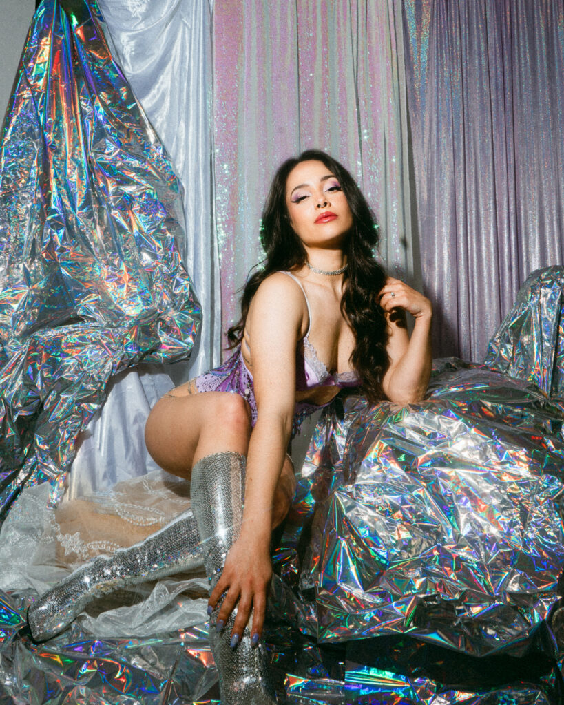 Burlesque performer Scarlett Snow poses in a sea of holographic fabric. She is wearing knee-high silver boots, and a purple slip.