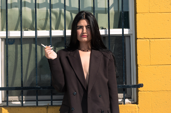 Lalya Said, a Romanian Afhgan poet, singer, and activist, stands in front of a window. The building behind her is yellow. Said is wearing a black sports coat and holding a cigarette in her hand.