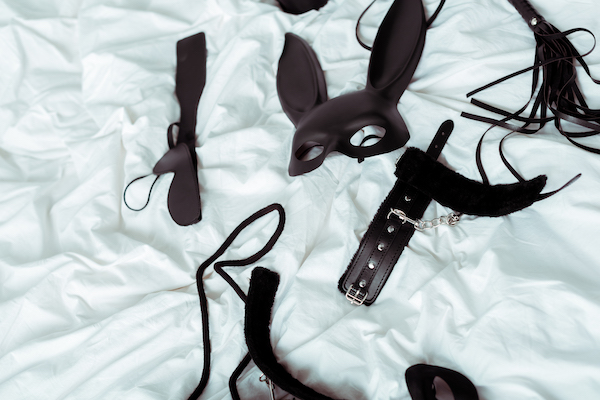 Various adult toys are laid out on a white sheet. They include a mask, cuffs, and a tawse for whipping.