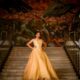 A young girl poses on a staircase in an ornate building. She is wearing a gold ball gown for her quinceńera.