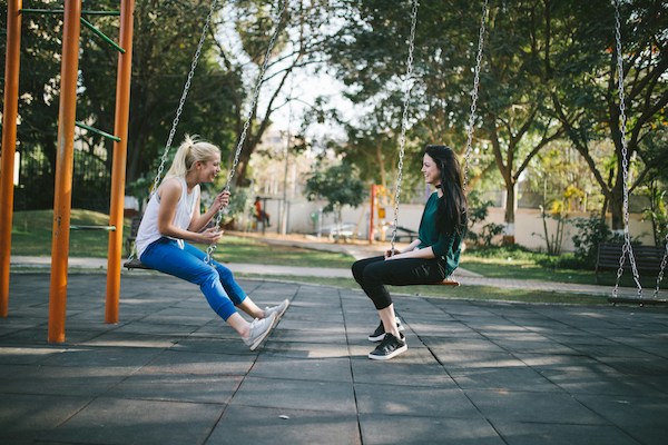 two women sit on playground swings, staring into each others eyes and smiling.