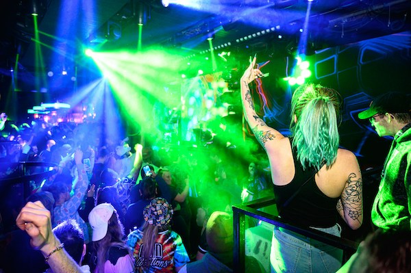 This photo shows a nightclub full of people. A neon green light overpowers blue and purple lights. A woman with jade green hair and tattoos stands on a balcony, moving her arm with the beat of the music. Below her, other people dance and move to the music.