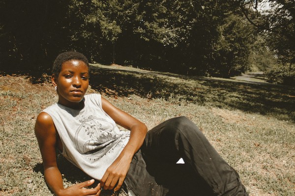 A Black person reclines on a grassy hill. Their medium-deep skin glistens in the sunlight. They have short cut coily hair. They are wearing a white graphic tank top and dark grey pants.
