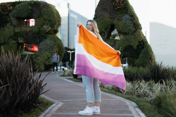 Digital creator and Instagram influencer Allyssa Leaton stands on a path in Buenos Aires, Argentina. She is holding a large lesbian flag which obscures a view of her body. Her head is visible and she smiles into the camera.