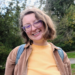 Writer Eryn Sunnolia poses outside against a backdrop of lush green trees. Eryn wears their hair in a light brownish blonde bob. She wears glasses with clear frames and a yellow top with a brown corduroy button-up. She smiles as she looks into the camera.