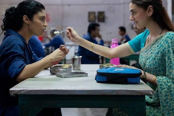 Two women sit across from one another at a table. One woman wears a dark blue shirt, the other a turquoise sari.