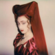 Musical artist Chappell Roan poses in a silky burgundy dress. A burgundy shawl (or veil) covers her hair. Roan's auburn ringlets peek out from under the shawl.