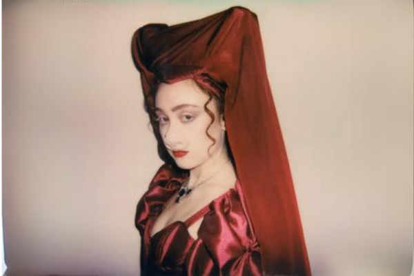 Musical artist Chappell Roan poses in a silky burgundy dress. A burgundy shawl (or veil) covers her hair. Roan's auburn ringlets peek out from under the shawl.