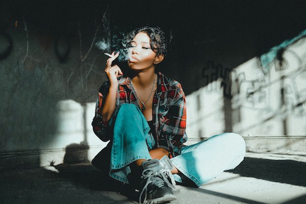 A woman sits on the floor in a dark room smoking a joint. She is wearing jeans, sneakers, and has wavy dark hair.