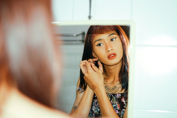A young Asian woman looks into a mirror as she puts on hoop earrings. She has long auburn hair with blunt bangs. She wears makeup, specifically a thick black cat-eye wing on each eye.