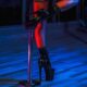 A woman bathed in blue light leans against a silver pole. She's wearing black leather platform stiletto boots.