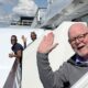 Three men stand outside of their cabin rooms on a cruise ship. They are smiling and waving.