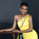 Shavana Clarke poses on a piano in a bright yellow dress. She is a beautiful Black woman with short curly hair.