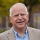 Tim Walz, a middle-aged white man with white hair, smiles kindly as he stands outside.