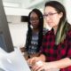 A Black woman and an Asian woman sit at a computer monitor, working together.