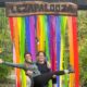 Two women smile and pose under a giant wooden sign that reads "Lezapalooza." Rainbow streamers hang from the sign, creating a bright background for the women.