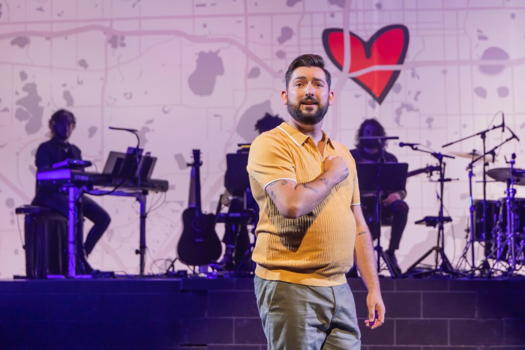 A man in a yellow shirt sings on stage with his hand on his heart. Behind him a band is playing.