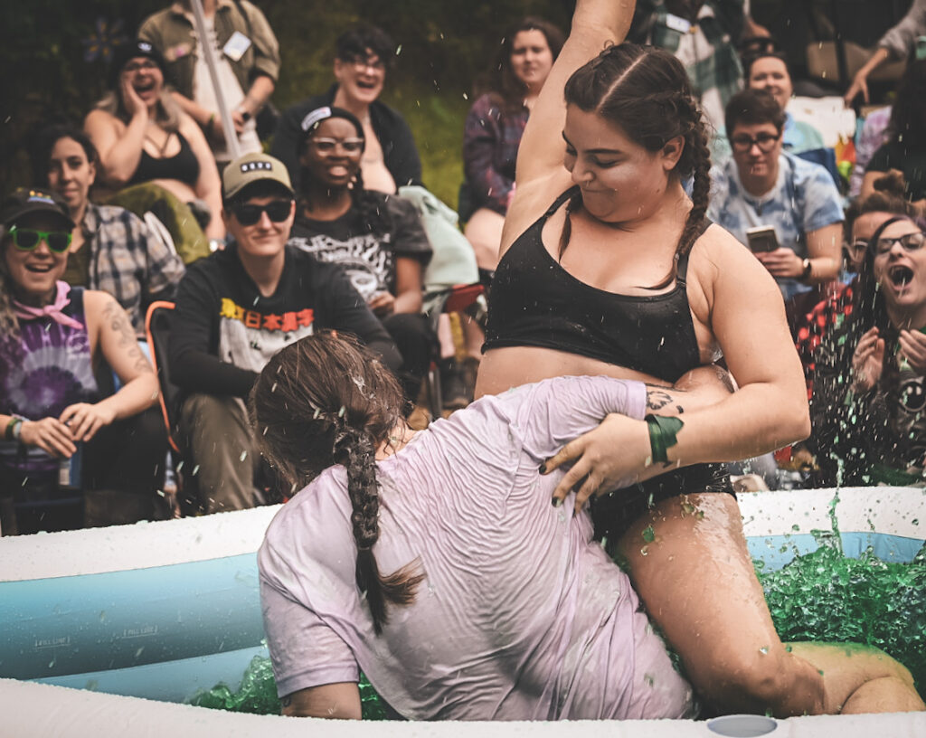 Two women—one in a sports bra and the other in a t-shirt—wrestle in a pool of Jell-O.