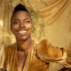 Shasheer Zamata, a beautiful Black woman, poses in a gold dress against a golden backdrop. Her skin is glowing.