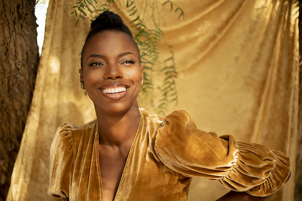 Shasheer Zamata, a beautiful Black woman, poses in a gold dress against a golden backdrop. Her skin is glowing.