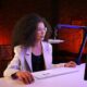 A young woman sits at a desk, looking a monitor as she prepares to speak into a microphone.