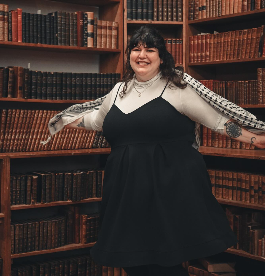 Author Hannah Moushabeck smiles in a library. She poses with her arms outstretched, holding a black and white Keffiyeh behind her like wings.