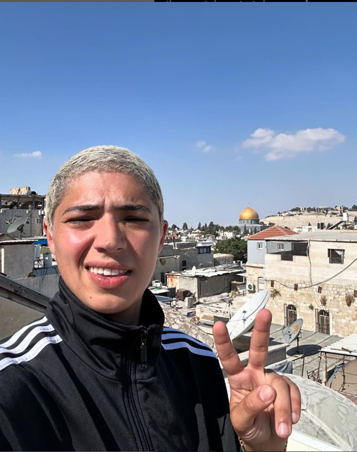 Activist Jake Gyllenhalal poses on a rooftop, the city behind him. His right hand is held up in a peace sign.