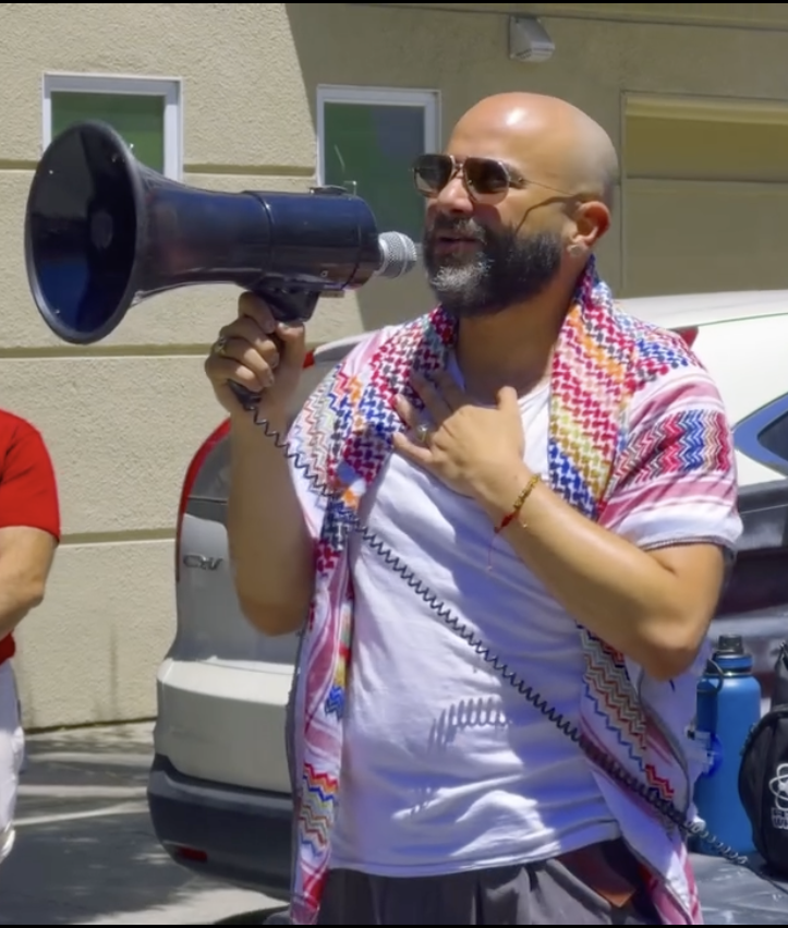 Drag artist Mama Ganuush speaks into a megaphone at Pride 2024. They are wearing a rainbow keffiyeh around their shoulders.