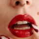 A woman with a septum piercing has bright red lips and sensually holds her manicured fingers up to her open lips.