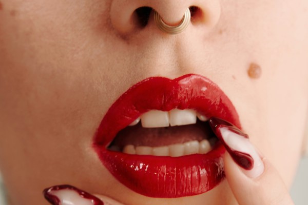 A woman with a septum piercing has bright red lips and sensually holds her manicured fingers up to her open lips.