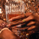 A Black woman's hand holds a glass of whiskey. She has gold rings on her fingers.