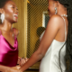 Two women in cocktail dresses hold hands and smile at one another.